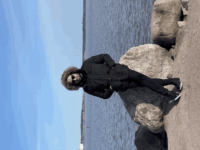a man in a black jacket with a furry hood stands on a rock near the water