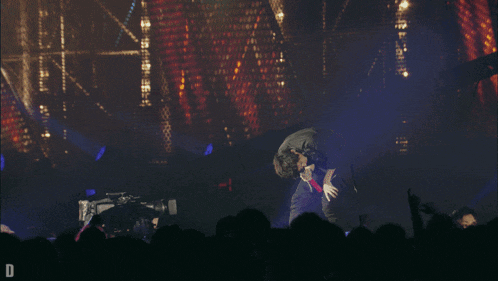 a man stands on a stage in front of a crowd with the letter d in the corner