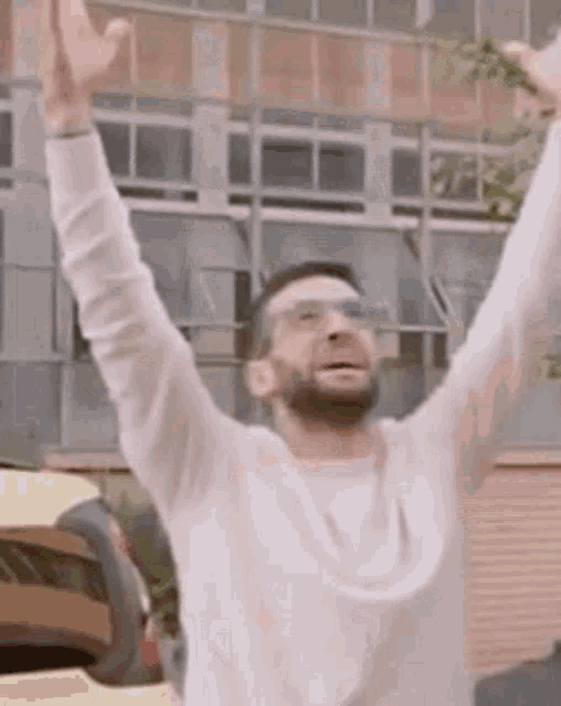 a man in a white sweater is raising his arms in the air in front of a building .