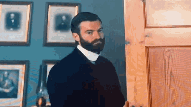 a man with a beard is standing in front of a door with pictures on the wall behind him