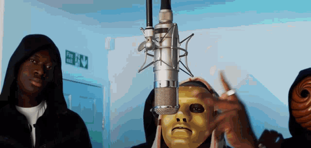 a man wearing a mask stands in front of a microphone with an exit sign on the wall behind him