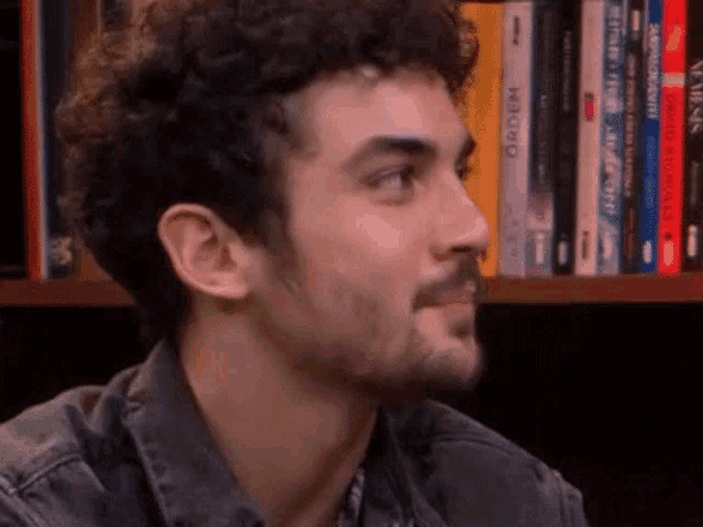 a man with curly hair and a beard is sitting in front of a bookshelf with books on it .