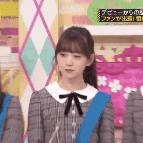 a girl in a plaid dress is standing in front of a microphone with a blue sash around her waist .