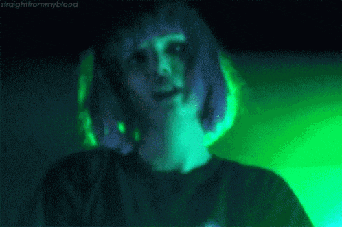 a person with purple hair is standing in front of a green light and covering their face with their hand .