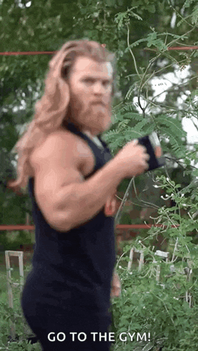 a man with long hair and a beard is standing in front of a tree and holding a dumbbell .