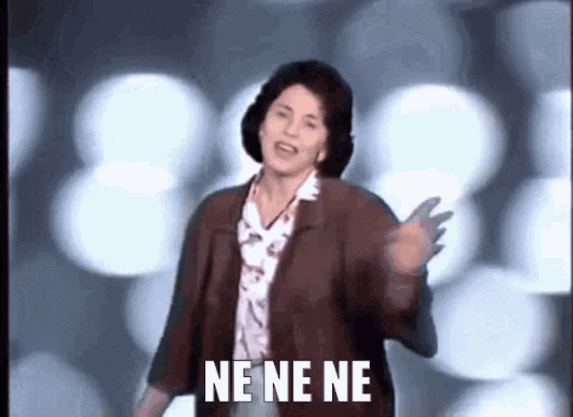 a woman in a brown jacket says ne ne ne in front of a blurry background