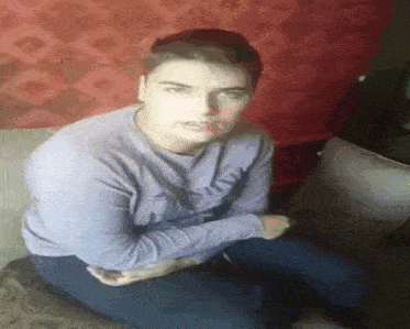 a young man in a purple shirt is sitting on a couch with his hands folded in front of him