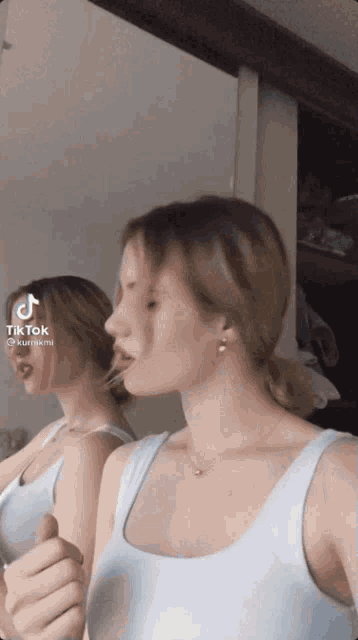 a woman in a white tank top is looking at herself in a mirror with tik tok written on the bottom right