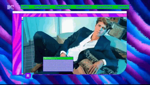 a man in a suit sits on a couch with a purple and blue background and a green r on the bottom right