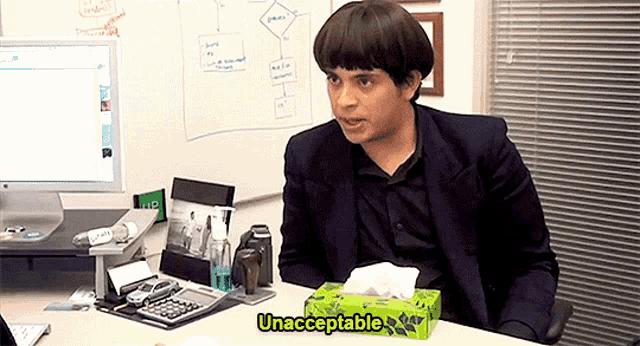 a man sitting at a desk with a box of kleenex next to him that says unacceptable on it