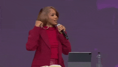 a woman in a red jacket is holding a microphone while standing in front of a laptop and a bible .