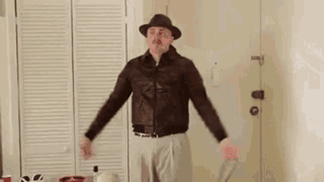 a man wearing a hat and a leather jacket is standing in a room with a suitcase .