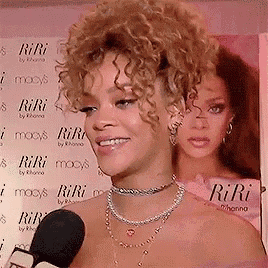 a woman with curly hair is talking into a microphone in front of a sign that says riri