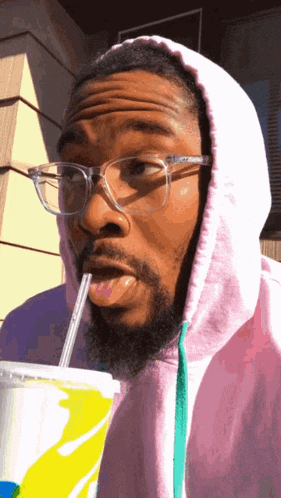 a man wearing glasses and a pink hoodie drinks from a cup with a straw