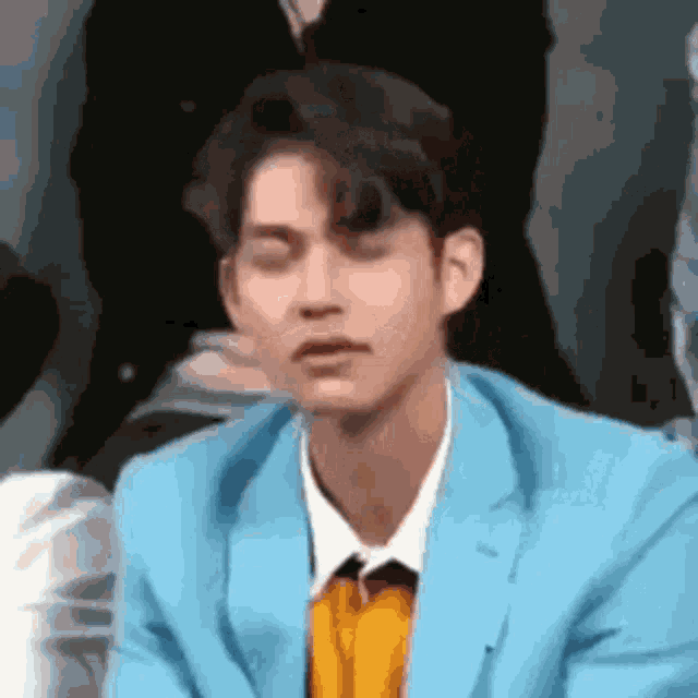 a man in a blue suit and yellow shirt is sitting in front of a group of people and making a funny face .