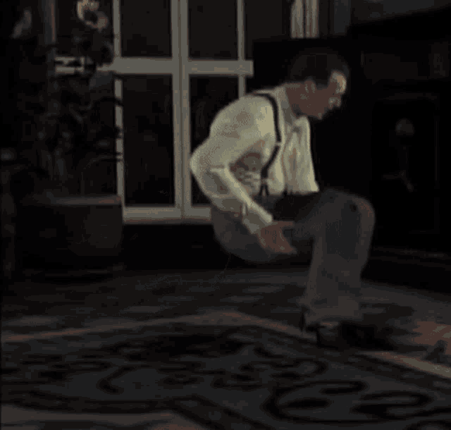 a man in suspenders is squatting on a rug in a dark room