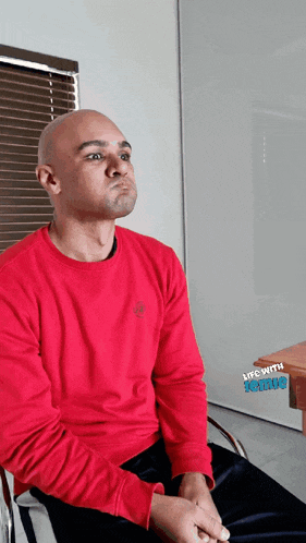a bald man wearing a red sweater is sitting in front of a window with blinds