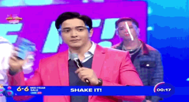 a man in a pink suit is holding a microphone in front of a banner that says shake it