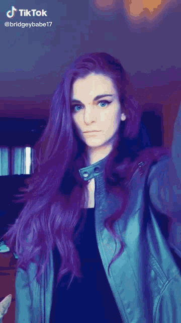 a woman with long purple hair is wearing a blue jacket and a black top .