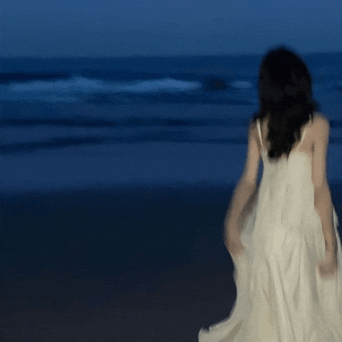 a woman in a white dress is walking on a beach at night
