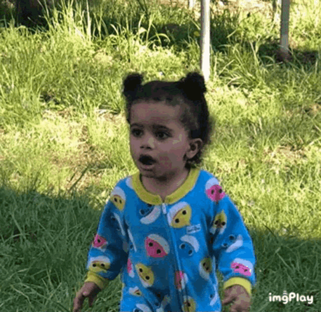 a little girl in a blue pajamas is standing in the grass making a funny face .