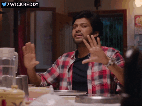 a man in a plaid shirt is sitting at a table with his hands outstretched in front of a sign that says 7wickreddy