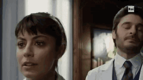 a man and a woman are standing next to each other in a room . the man is wearing a white coat and tie .