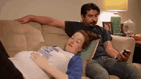 a man and a pregnant woman are sitting on a couch . the woman is sleeping on the man 's lap .