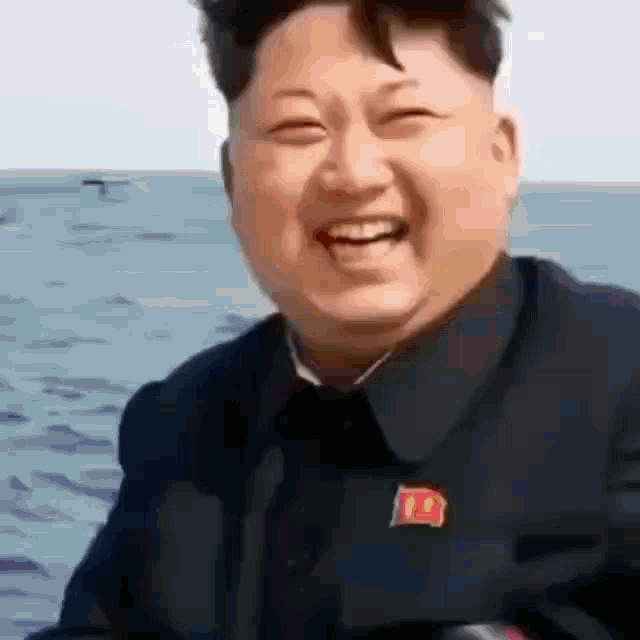 a man in a black suit with a red badge on his chest smiles in front of the ocean