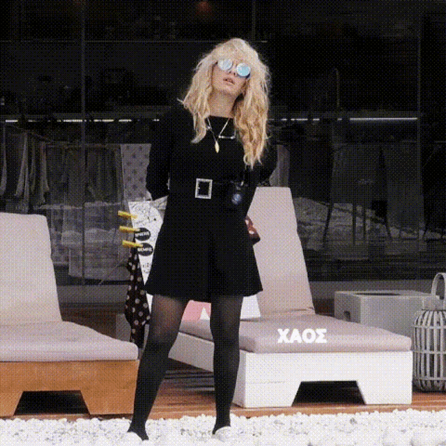 a woman wearing sunglasses and a black dress is standing in front of a chair with the word xaos on it