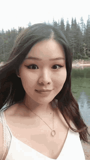 a woman wearing a white tank top and a necklace stands in front of a lake with trees in the background