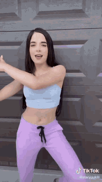 a woman in a blue crop top and purple pants is dancing in front of a garage door