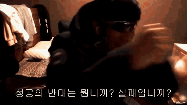 a man wearing sunglasses is sitting in a dark room with korean writing on the bottom of the screen