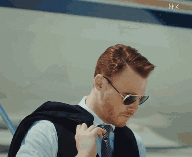 a man wearing sunglasses and a suit is standing in front of an airplane with hk written on the bottom