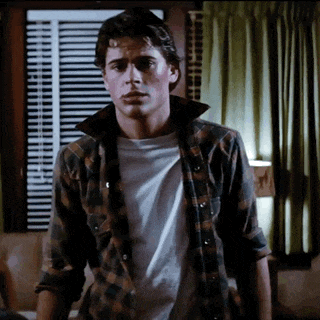 a young man wearing a plaid shirt and a white t-shirt stands in front of a window