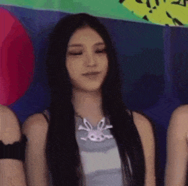 a woman with long black hair and a bunny necklace is sitting in a room with other people .