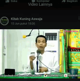 a man is speaking into a microphone and holding a cellphone in front of a screen that says kitab kuning aswaja