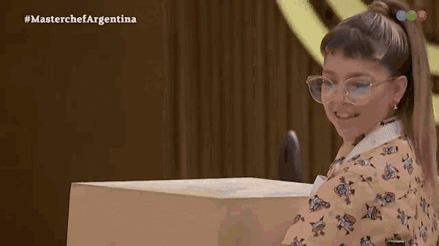 a little girl wearing glasses is sitting at a table in front of a screen that says masterchef argentina on it