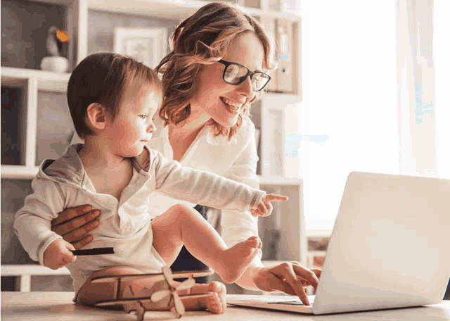 a woman with glasses holds a baby while looking at a laptop