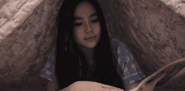 a girl in a blue pajama top is reading a book under a blanket
