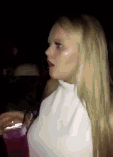 a woman with long blonde hair is sitting in a dark room holding a cup of drink .