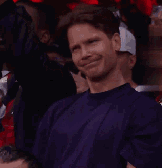 a man in a purple shirt is sitting in a crowd of people and making a funny face .
