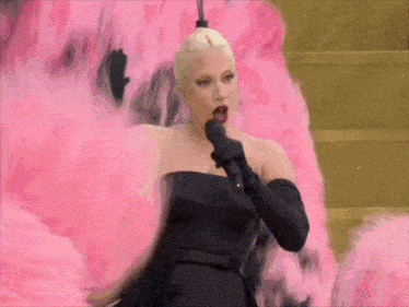 a woman in a black dress is singing into a microphone while surrounded by pink feathers .