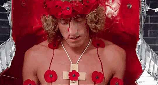 a shirtless man with a cross necklace and red flowers on his chest is laying in a coffin .