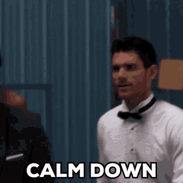 a man in a tuxedo is standing in front of a blue wall and says calm down .
