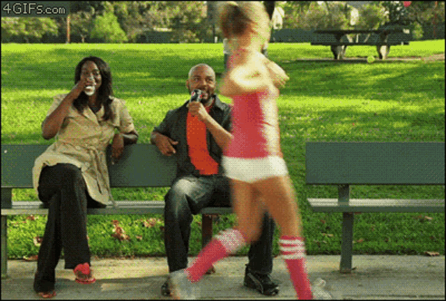 a woman sitting on a park bench drinking a bottle of water while a girl in pink shorts runs past her