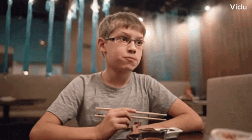 a boy wearing glasses sits at a table with chopsticks in front of a vidu banner