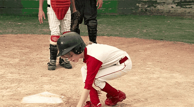 a young boy in a red and white baseball uniform squats on the base