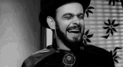 a black and white photo of a man with a beard wearing a top hat and cape laughing .