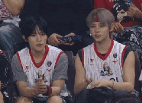 two young men in basketball uniforms are sitting in the stands watching a game .